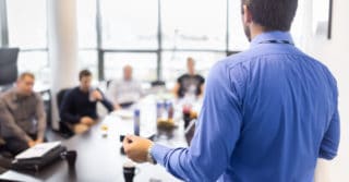 A man stands with his back to the camera. He talks to a group of people sat around a large table.