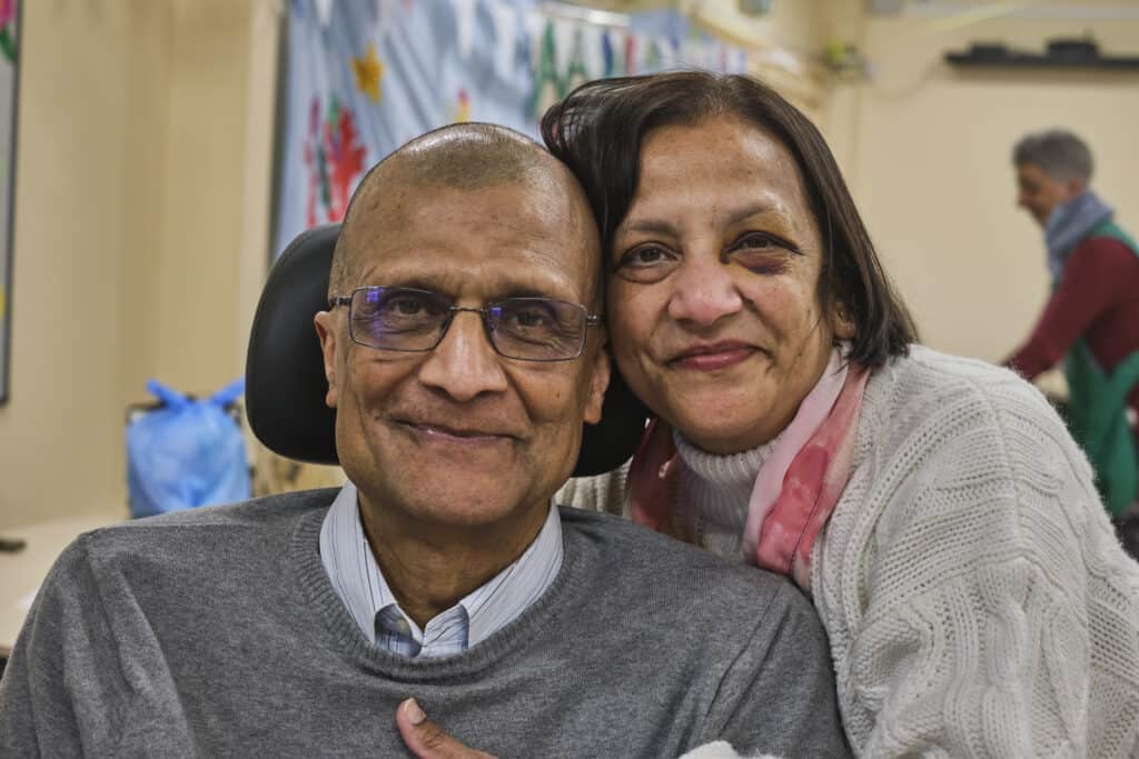 A woman with her arms around a seated man, both are smiling.