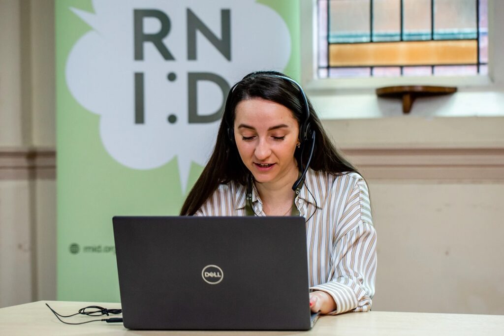 A woman sat at a laptop and wearing a headpiece, speaking with a caller down the phone. She's smiling, and in the background is an RNID banner.