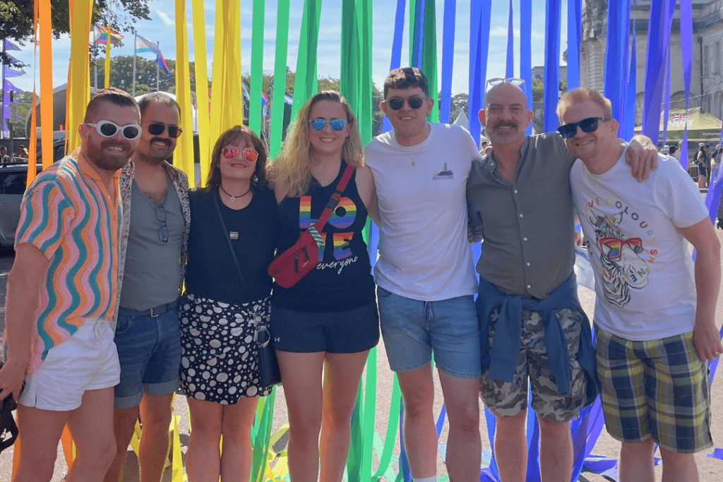 A photograph of Chris with his friends, wearing sunglasses and shorts, standing outside in front of some colourful streamers. 