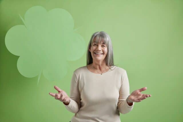 A woman with long grey hair sits smiling in front of a green background.