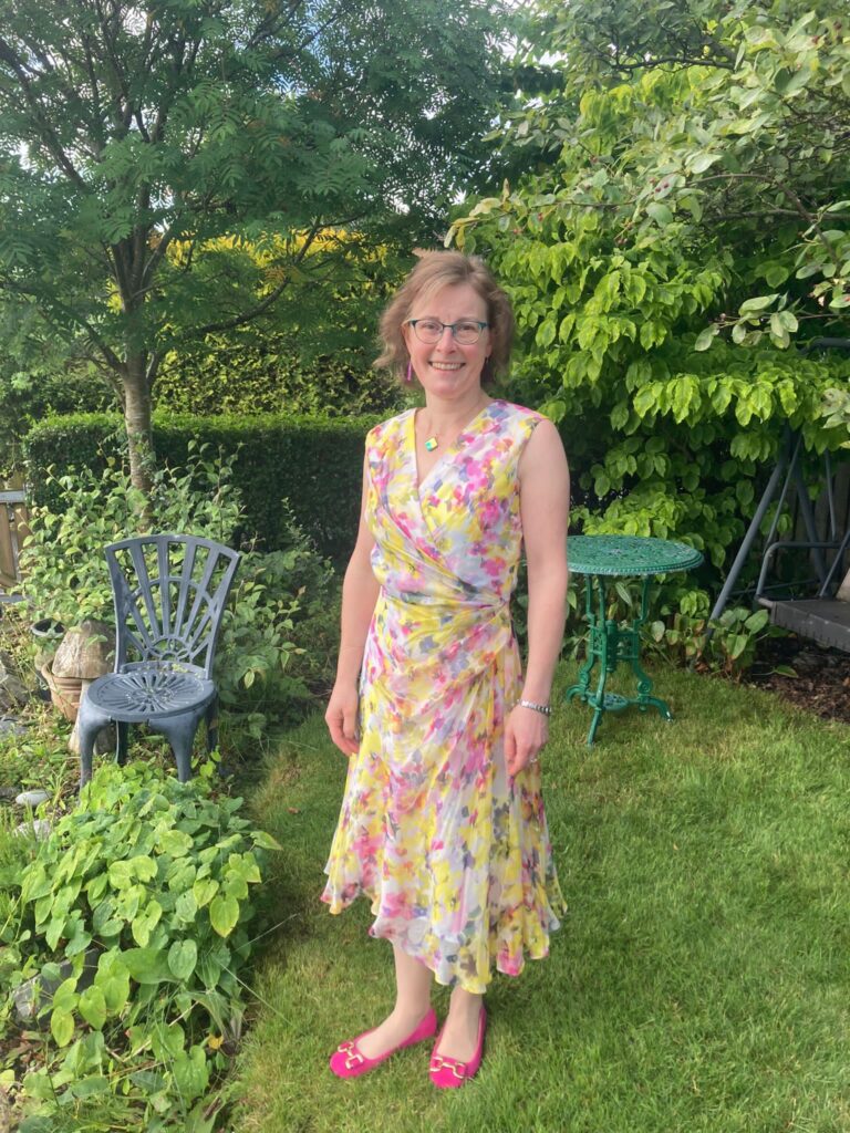 RNID volunteer Clare from Peebles, standing in a garden and wearing a summer dress, smiling at the camera.