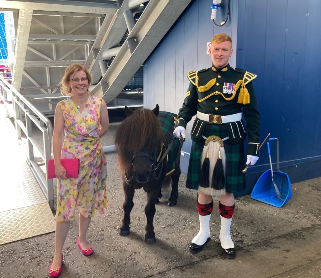 RNID volunteer Clare from Peebles standing in a summer dress with a man in a military uniform and kilt, with a small horse. 