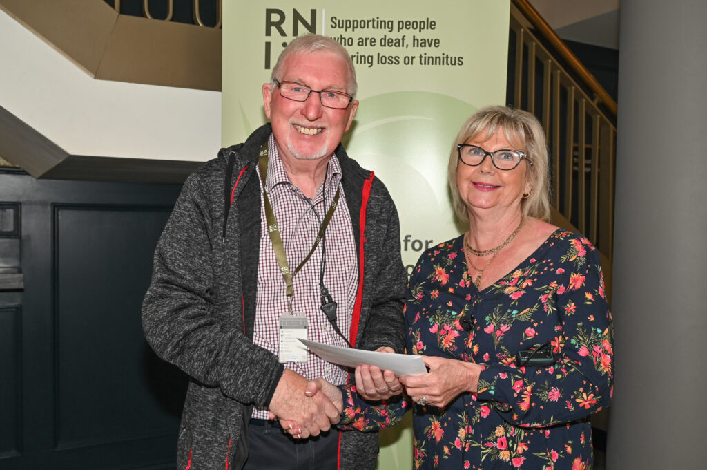 Gerry, an older man with a goatee, with Cilla Mullan, RNID Manager in Northern Ireland: a woman smiling with glasses, shaking hands and holding a certificate between them. 