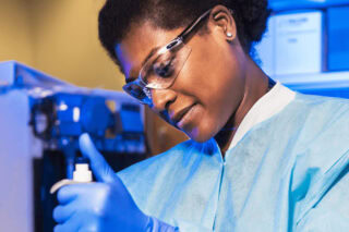 A woman in a laboratory wears a lab coat, gloves and protective goggles.