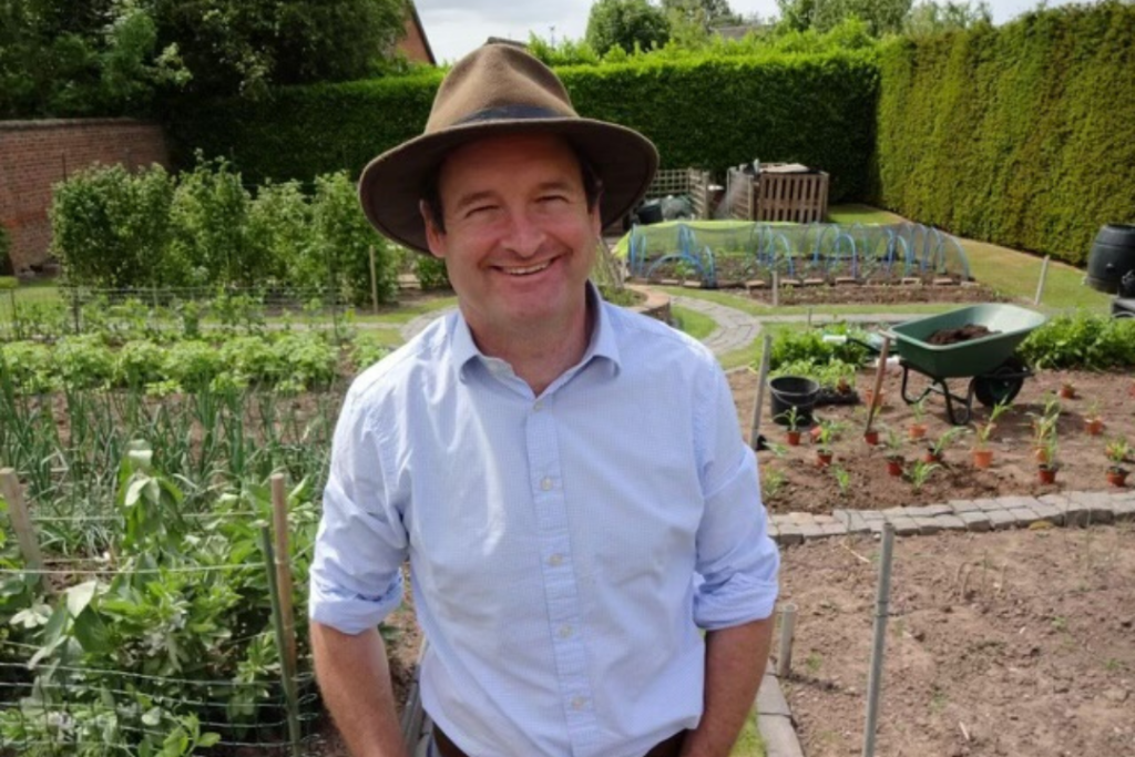 Hugh, a man in a shirt and sunhat, stands outside in a garden and smiles at the camera. 