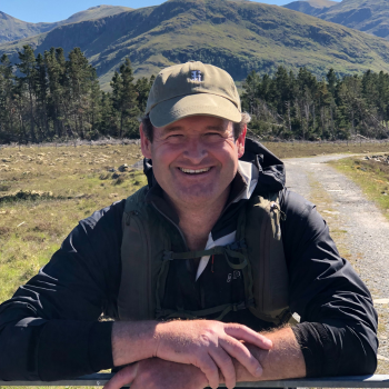 Hugh, outside in front of Scottish mountains, leaning on a fence post and smiling at the camera. He wears a sun cap and a hiking rucksack. 