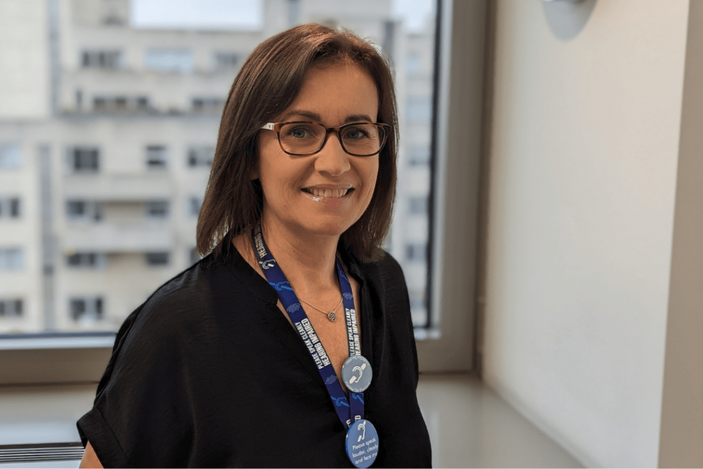 A photo of Sandra, a woman with mid-length brown hair and glasses, wearing a hearing loss awareness lanyard in the workspace.