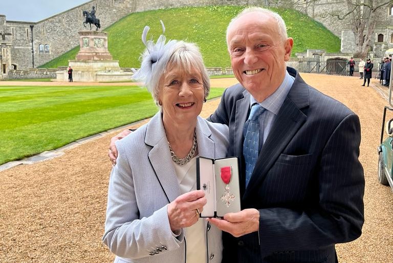 Photograph of Brian and his wife Sandra, standing outside. They hold Brian's MBE