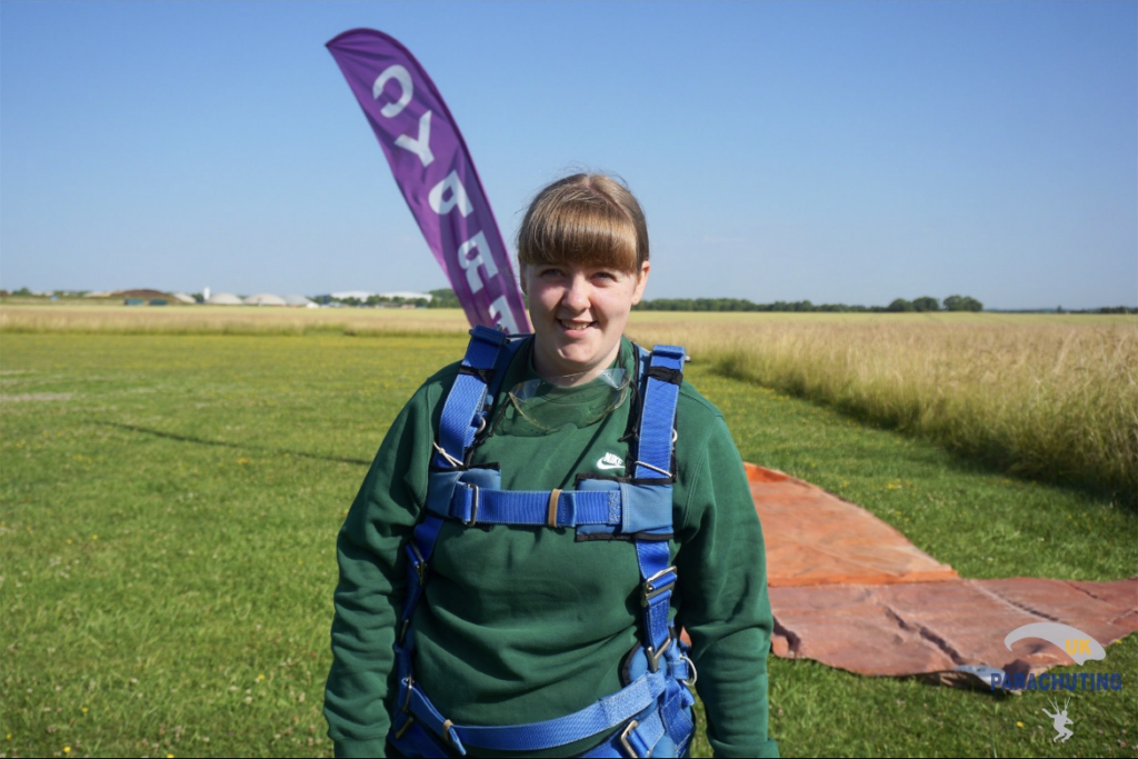 A photograph of Ellie, a teenager with long brown hair in a ponytail and jumper, wearing a parachute and standing in a large flat field. She's smiling at the camera. 
