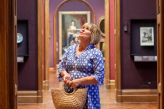 A woman stands looking at art in the National Portrait Gallery. She holds a straw bag and wears a blue dress.
