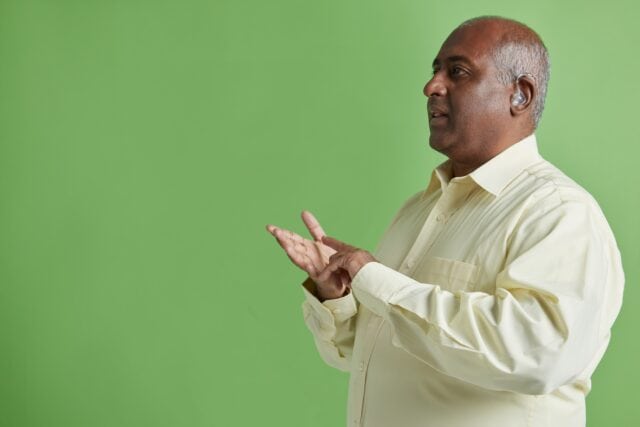 A man wearing a light yellow shirt using sign language. He is a hearing aid user.