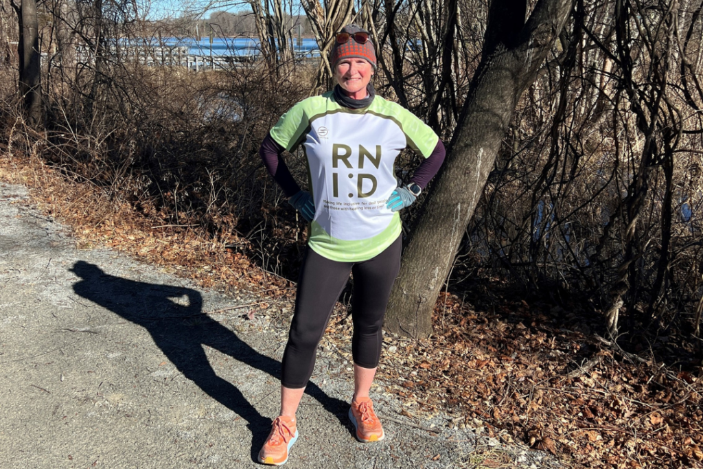 Deborah on a training run, looking out for bears! Deborah stands on a trail in front of trees and a lake, wearing a hat and an RNID running shirt. 