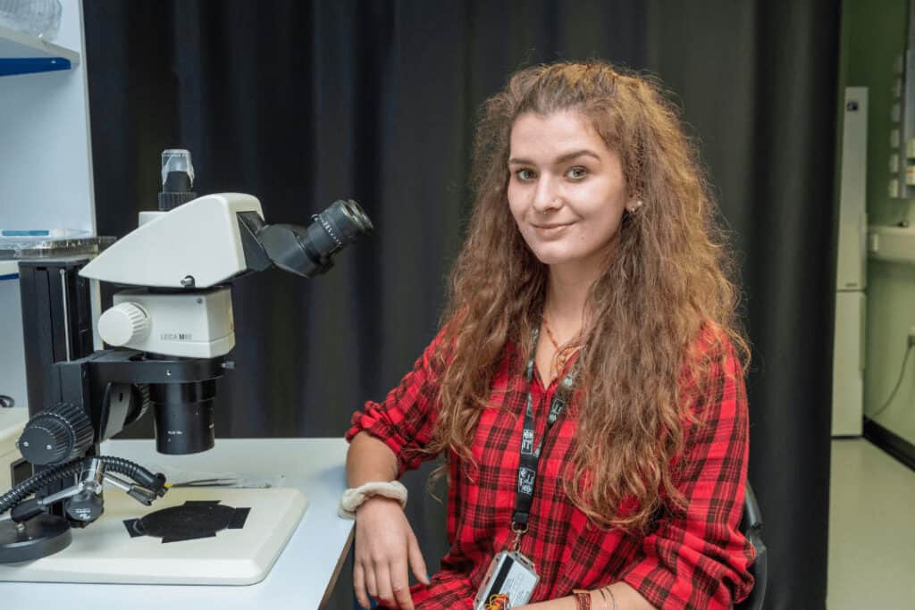 Alice Zanella, a PhD student funded by RNID. Alice sits at a microscope and smiles at the camera.