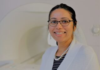 Professor Fatima Husain in a lab. She wears a white cardigan and pearls and is smiling at the camera.