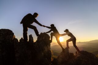 Three people help each other climb the rocky ledge of a mountain as the sun sets.
