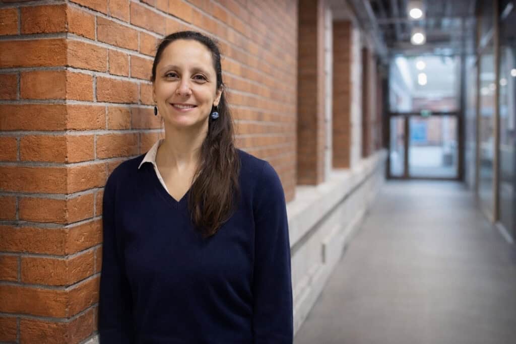 A smiling woman with brown hair tied in a ponytail stands beside a brick wall and smiles.